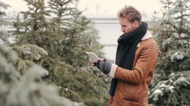 Şık giyinmiş yakışıklı bir adam not defteri kullanıyor ve markette noel ağacını seçiyor.