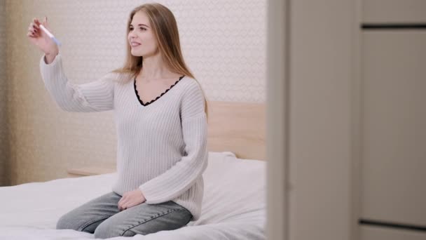 Молода леді відчуває себе щасливою, що завагітніла, що вона дізналася від тесту на вагітність, який вона тримає, сидячи на ліжку — стокове відео