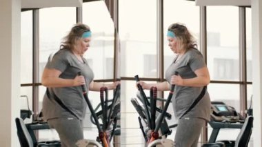 Orta yaşlı şişman kadın spor salonunda elips makinesi kullanıyor.