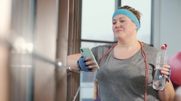 Mulher com excesso de peso no ginásio moderno na janela segurando smartphone — Vídeo de Stock