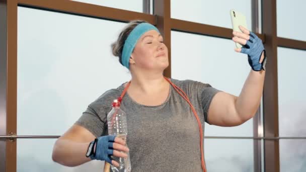Übergewichtige Frau in modernem Fitnessstudio am Fenster mit Smartphone — Stockvideo
