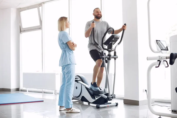 Ασθενής κάνει άσκηση με ποδήλατο σπιν στο γυμναστήριο με θεραπευτή — Φωτογραφία Αρχείου