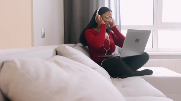 Ung afrikansk kvinna i hörlurar med bärbar dator som arbetar hemma — Stockvideo