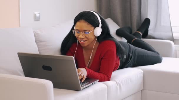 Ung afrikansk kvinna i hörlurar med bärbar dator som arbetar hemma — Stockvideo
