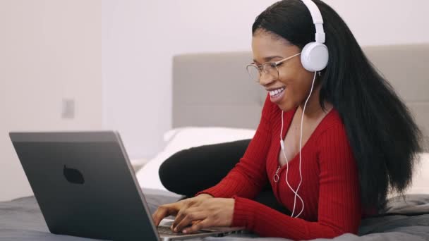 Ung afrikansk amerikansk kvinna njuter lyssna på musik på laptop när du ligger på sängen — Stockvideo