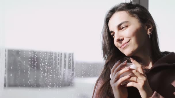 Молодая женщина в капюшоне возле окна в дождливый день — стоковое видео