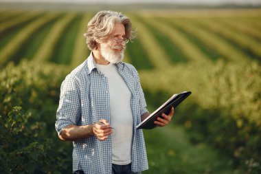 Tişörtlü yaşlı çiftçi elinde defterle sahada duruyor.