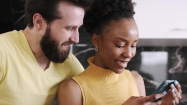 Çok ırklı bir çift mutfaktaki akıllı telefondan bir şey kontrol ediyor.