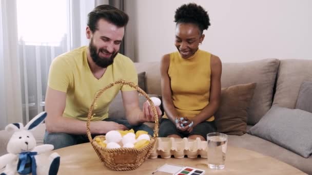 Чорна жінка і європейський чоловік малюють разом пасхальні яйця. — стокове відео
