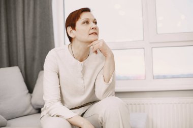 Olgun, şık bir kadın. Evdeki koltukta oturuyor.