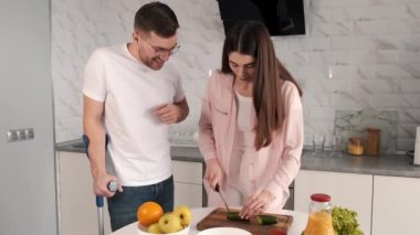 Kadın, engelli kocayı mutfak masasında tekerlekli sandalyede oturmaya teşvik eder.