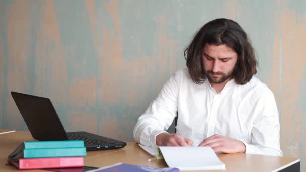 Młody beraded człowiek z długimi włosami w białej koszuli pracuje w biurze z papierami i laptopem — Wideo stockowe