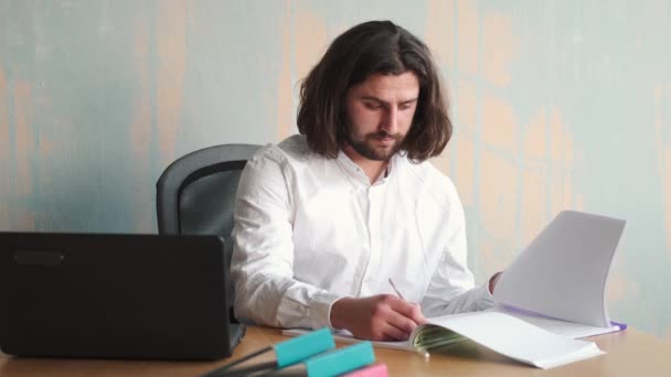 Jeune homme beradé aux cheveux longs en chemise blanche travaillant au bureau avec des papiers et un ordinateur portable — Video