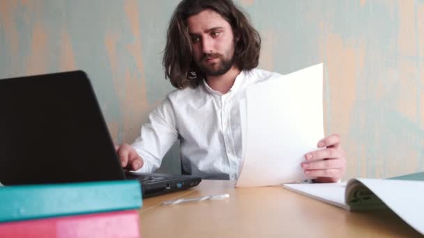 Jeune homme beradé aux cheveux longs en chemise blanche travaillant au bureau avec des papiers et un ordinateur portable — Video