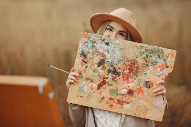 Kahverengi ceketli bir kadın tarlada resim yapıyor.