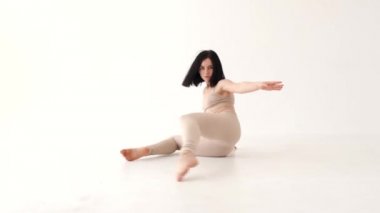 Bej kostümlü bir kadın beyaz arka planda dans ediyor.