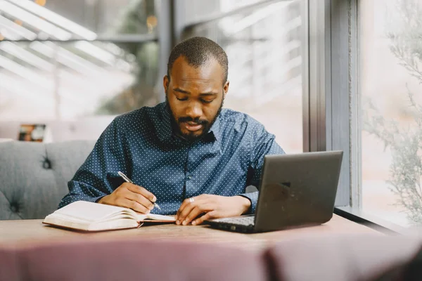 Homem sentado em um café e trabalhando atrás de um laptop, escrevendo em um caderno. — Fotografia de Stock