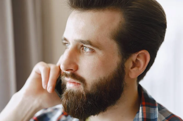 Чоловік з бородою стоїть біля вікна з телефоном — стокове фото