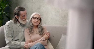 Oturma odasındaki kanepede oturan gündelik kıyafetli yaşlı çift.