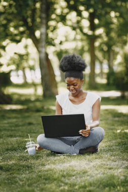 Elinde dizüstü bilgisayarla çimlerin üzerinde oturan gülümseyen Afro-Amerikalı bir kızın portresi.