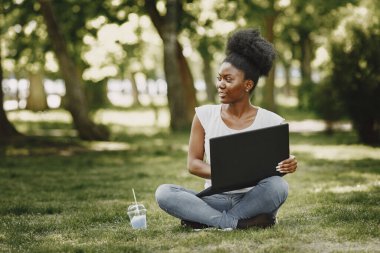 Elinde dizüstü bilgisayarla çimlerin üzerinde oturan gülümseyen Afro-Amerikalı bir kızın portresi.