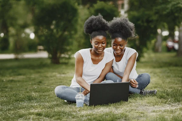 Jovens irmãs gêmeas afro-americanas sentadas em uma grama em um parque e usando um laptop — Fotografia de Stock