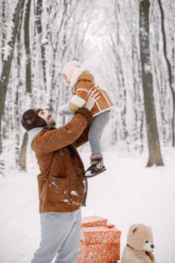 Baba küçük kızını elinde tutuyor ve kar tatilinde ormanda eğleniyor. Sakallı adam ve küçük kız kalın kıyafetler giyiyorlar. Hediye kutuları ve yanında duran büyük ayı oyuncakları..