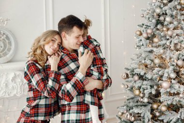 Sarışın anne, esmer baba ve Noel ağacının yanında duran küçük kız. Ekose pijama giyen bir kadın ve kız. Noel 'i birlikte kutlayan aile.