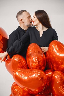 Yıldönümünde fotoğraf çektirmek için poz veren romantik bir çift. Adamın karısını kucakladığı ve elinde kalp şeklinde kırmızı balonlar tuttuğu bir stüdyo fotoğrafı. Esmer kadın ve siyah elbiseli sakallı adam..