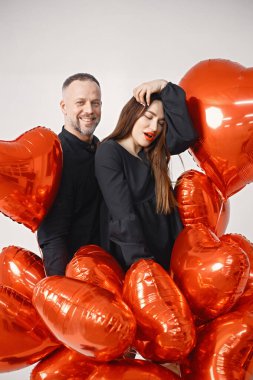 Yıldönümünde fotoğraf çektirmek için poz veren romantik bir çift. Adamın karısını kucakladığı ve elinde kalp şeklinde kırmızı balonlar tuttuğu bir stüdyo fotoğrafı. Esmer kadın ve siyah elbiseli sakallı adam..