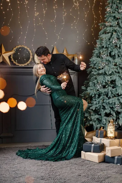 Genç bir kadın ve bir adam yılbaşı ağacının yanında duruyorlar ve ellerinde maytap var. Mutlu bir aile Noel 'i kutluyor. Yeşil elbiseli sarışın kadın ve siyah tişörtlü sakallı adam..