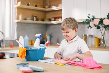 Sarışın çocuk mutfağı temizliyor. Beyaz çocuk annesine ev işlerinde yardım ediyor. Masanın üzerinde farklı halılar ve deterjanlar olan mavi bir kova..
