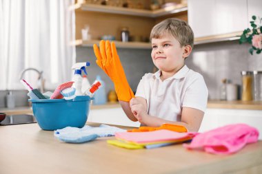 Sarışın çocuk mutfağı temizliyor ve turuncu eldiven giyiyor. Beyaz çocuk annesine ev işlerinde yardım ediyor. Masanın üzerinde farklı halılar ve deterjanlar olan mavi bir kova..