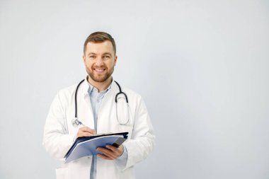 Dosyasıyla ayakta duran erkek doktor, beyaz arka planda izole edilmiş. Elinde dosya tutan bir erkek doktorun portresi var. Beyaz tıbbi önlük giyen ve steteskobu olan bir adam..