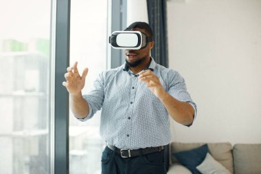 Black business man standing in office near big windows. Bearded man wearing virtual reality glasses. Man wearing blue shirt.
