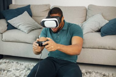 Black man in vr glasses, playing video games with virtual reality headset. Man wearing blue t-shirt. Man in virtual reality glasses sitting at home near coach.