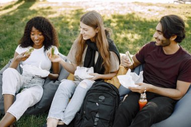 Friends hanging in the green park, relaxing. Boy and girls holding a bottles of juice. African girl wearing white clothes, boy wearing brown t-shirt and black jeans.