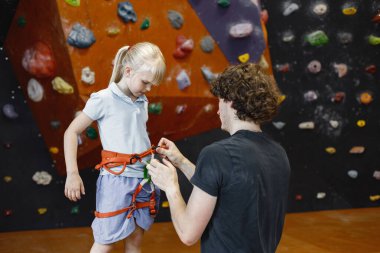 Eğitmen, küçük bir kızın içerideki kaya tırmanış merkezinde emniyet kemeri takmasına yardım ediyor. Küçük kız, çocuk eğlence merkezinde kaya tırmanışı yapıyor. Şort ve tişört giyen sarışın beyaz kız..