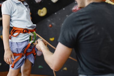 Küçük bir kızın içerideki kaya tırmanış merkezinde emniyet kemeri takmasına yardım eden bir eğitmenin göbeğinde. Küçük kız, çocuk eğlence merkezinde kaya tırmanışı yapıyor. Kız şort ve tişört giyiyor..