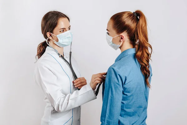 Doktor, beyaz bir arka planda steteskopla maskeli bir kadının sağlığını kontrol ediyor.