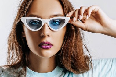 Güzel, genç, esmer, yeşil gözlü, moda makyajlı, mor dudaklı, izole gri arka planda güzel bir kız. Mavi güneş gözlüklü kadın ciddi ciddi kameraya bakıyor. Güzellik konsepti.