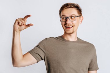 Gözlüklü yakışıklı esmer adam parmaklarını küçük gösteriyor ve beyaz izole edilmiş arka planda gülümsüyor.
