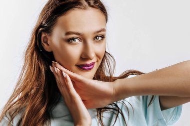 Güzel bir genç kız gülümser, hayal kurar ve ellerini yüzünün yakınında beyaz, ıssız, gri bir arka planda tutar. Reklam için yer var. Mor dudaklı, modaya uygun bir kadın..