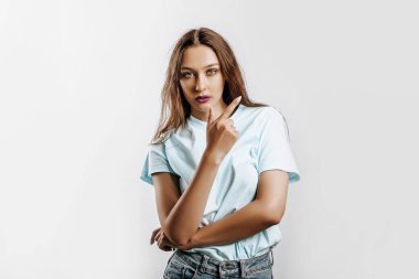 Genç ve güzel bir kız, beyaz izole bir arka planda parmağını yana doğru gösteriyor. Bir kadın bir fikri işaret eder, reklam için bir yer. Mor dudaklı, ciddi bir kız..