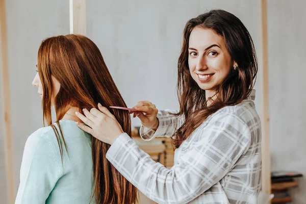 Two girls braid their hair at the window. Woman makes a braid to her friend. Hair weaving hairstyles. Girlfriend braids her hands with ringlets. Hair care