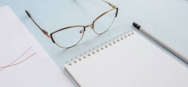 Ofis kavramı. Çalışma alanı gözlüklü, defterli ve kalemli. Boşluğu beyaz ve mavi olarak kopyala.