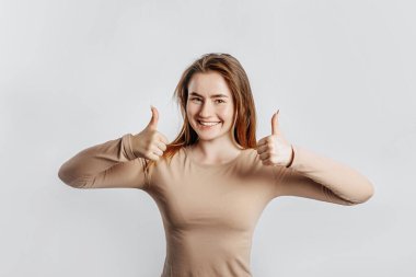 Güzel genç kız gülümsüyor ve beyaz izole bir arkaplanda baş parmak işareti yapıyor.