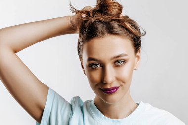 Güzel esmer bir kız saçını tutup kendi saç stilini yapar. Uzun saçlı genç bir manken, gri arka planda mor dudaklı bir moda makyajı. Kadın gülümser ve kameraya bakar..