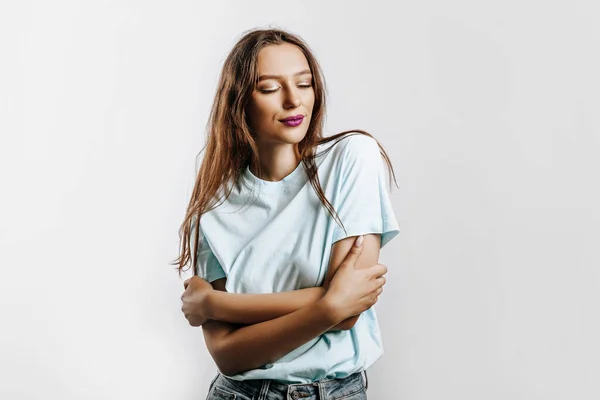 The woman hugs herself with closed eyes. Beautiful young brunette girl with green eyes with fashion makeup with purple lips on an isolated gray background. Kind cute model