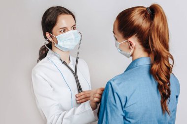 Doktor, beyaz bir arka planda steteskopla maskeli bir kadının sağlığını kontrol ediyor.